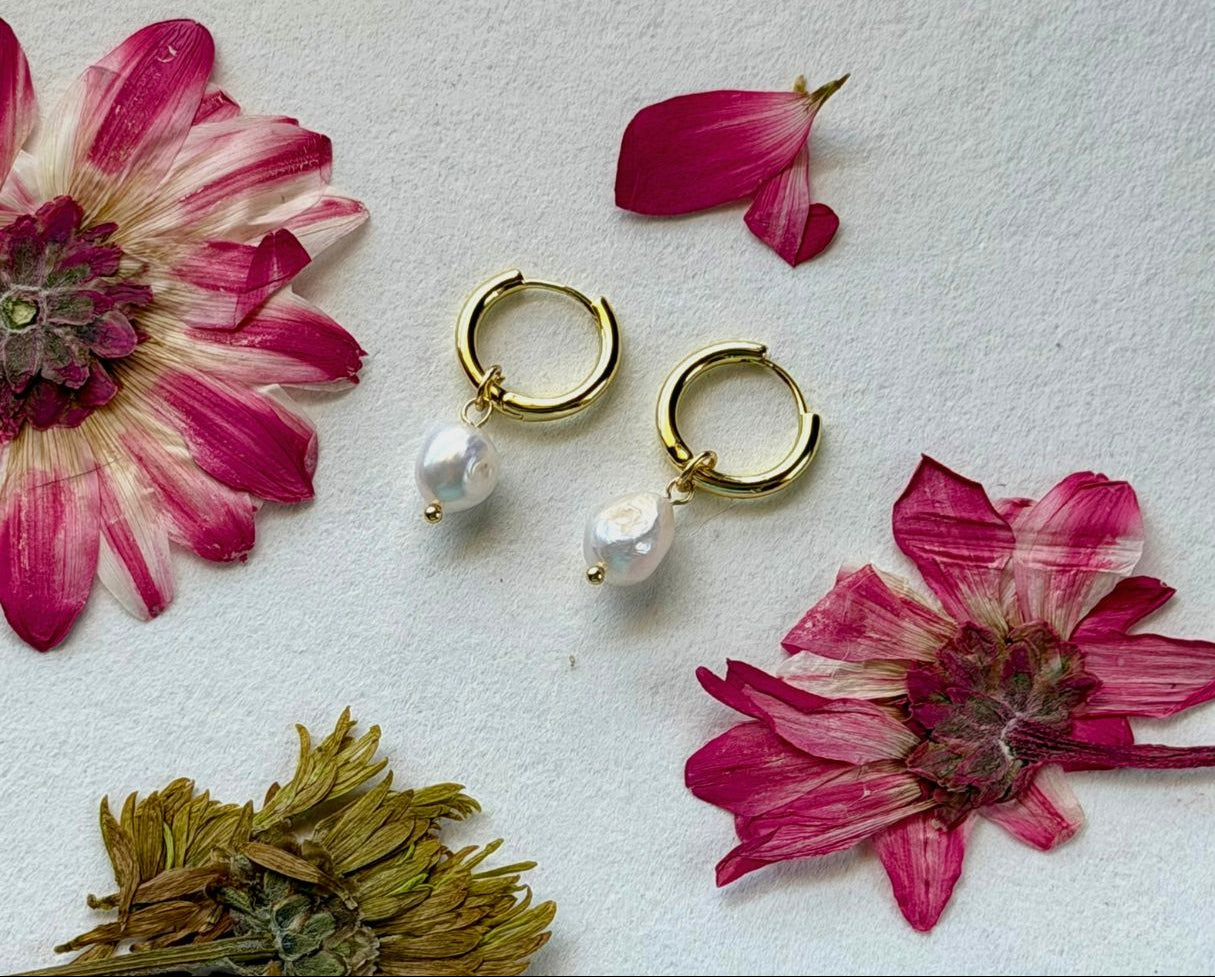 Gold hoop earrings with pearl beads on a white surface with pink flowers.