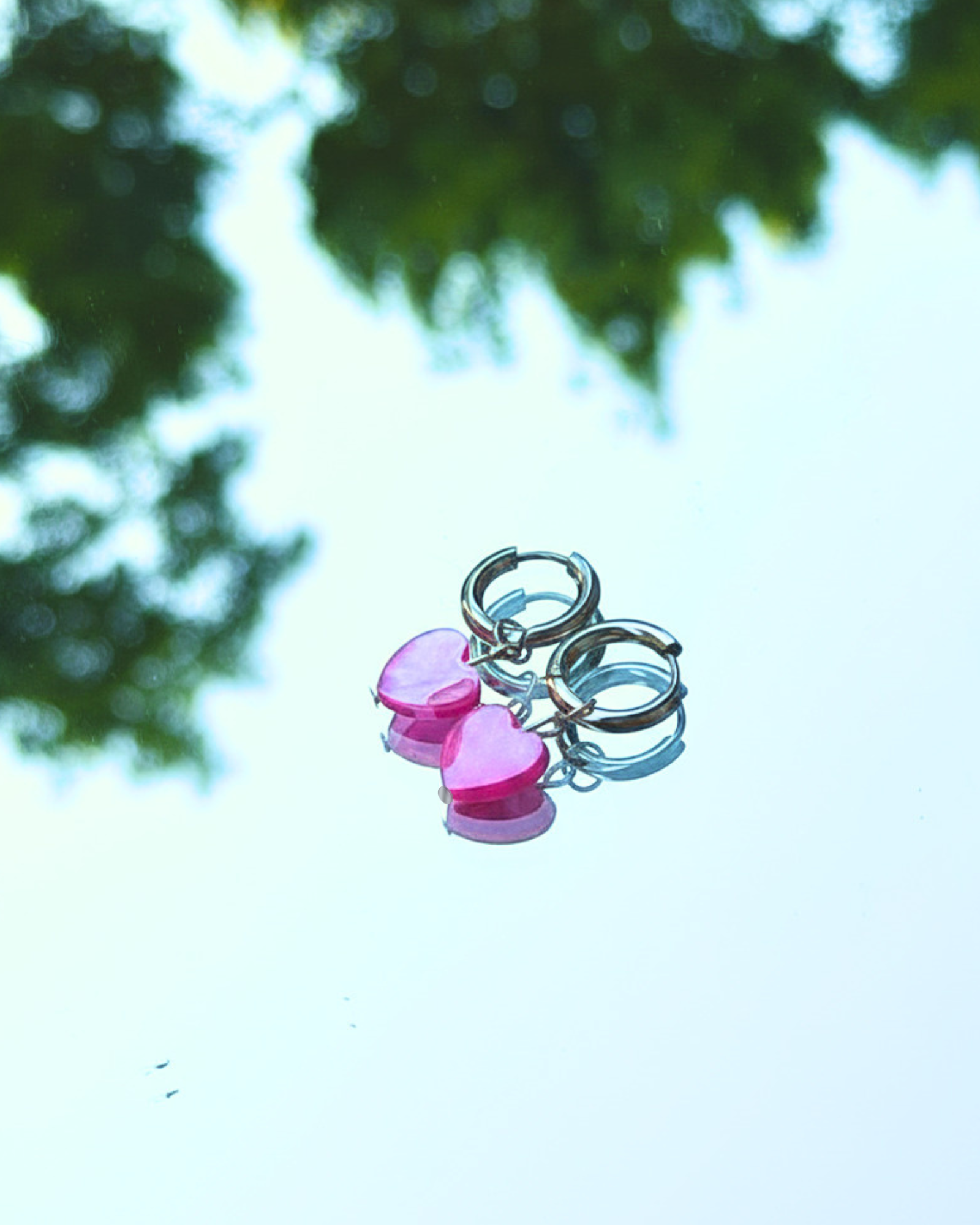 Two pink heart-shaped natural seashell dyed charms on silver 14mm hoop earrings against a blurred natural background