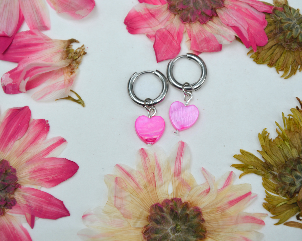 Silver hoop earrings with pink heart-shaped pendants on a white background with pressed flowers.